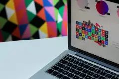 Ein Notebook, auf dem ein Grafikprogramm geöffnet ist, das Logos verschiedener Social Media-Plattformen zeigt. Im Hintergrund sind bunte Dreiecke.