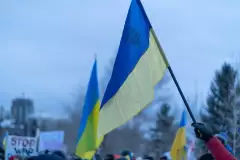 Ukrainische Flagge in der Hand einer protestierenden Person, im Hintergrund weitere Flaggen und Protestplakate