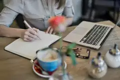Person schreibt an einem Tisch mit Laptop in ein Notizbuch