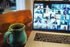 Ansicht eines Laptops bei einer Online-Konferenz (viele Gesichter), daneben eine Tasse.