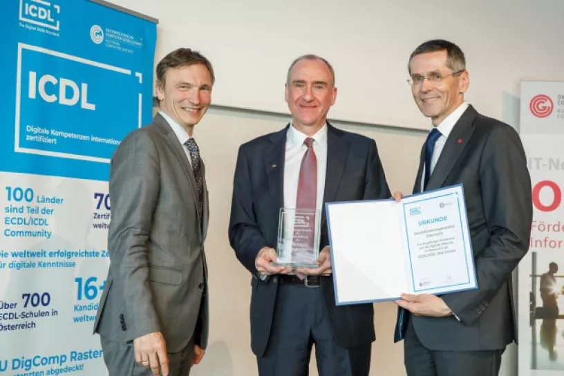Drei lächelnde Männer bei einer Preisverleihung, in der Mitte der Preisträger