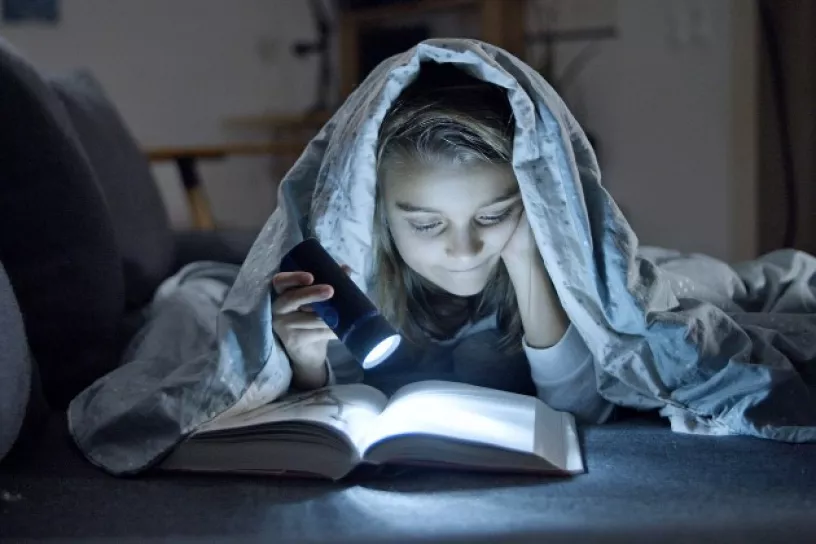 Kind liest mit einer Taschenlampe unter der Bettdecke ein Buch