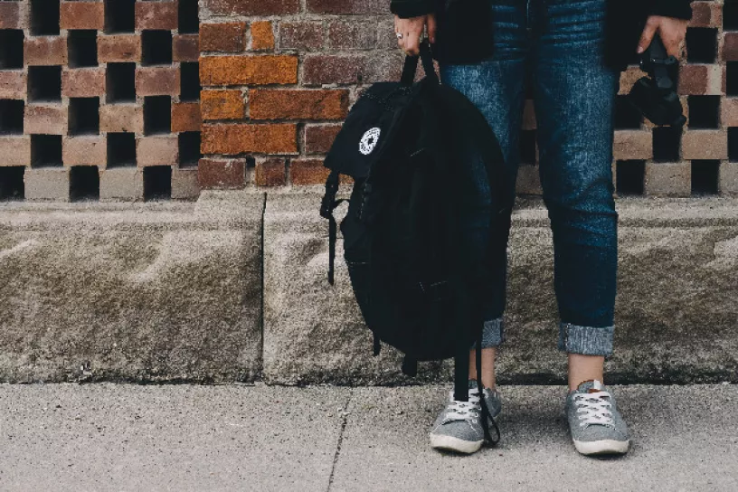 Eine Person steht mit einem Rucksack in der Hand vor einer braunen Wand