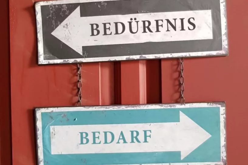 Wegweiser in verschiedene Richtung, darauf steht „Bedürfnis“ und „Bedarf“