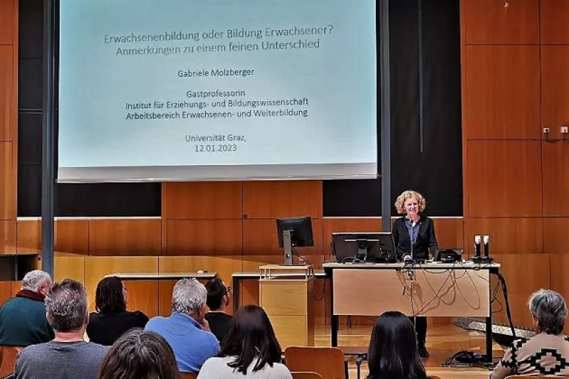 Vortragende an einem Pult vor Publikum, hinter ihr eine Leinwand mit Präsentation