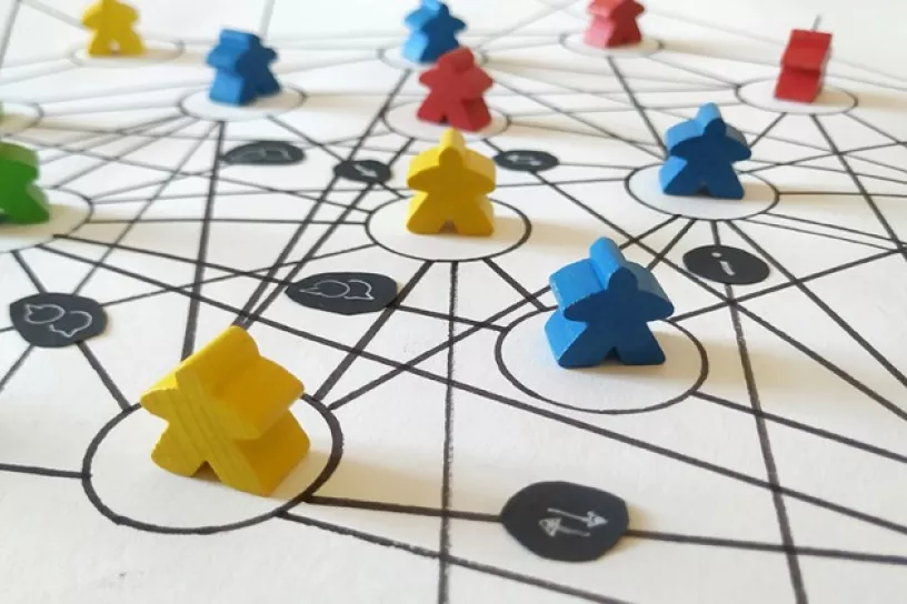 Spielfiguren stehen auf einem Papier, auf dem ein Netz gezeichnet ist, dazwischen sind Kommunikationssymbole dargestellt wie z.B. Sprechblasen, Rufzeichen und Pfeile