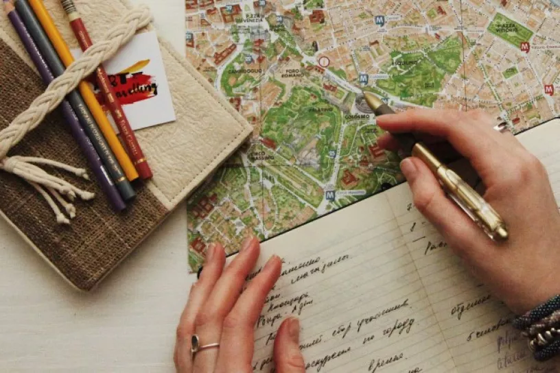 Ein Stadtplan, Stifte und ein Notizbuch; auf dem Notizbuch liegen Hände, eine hält einen Stift.